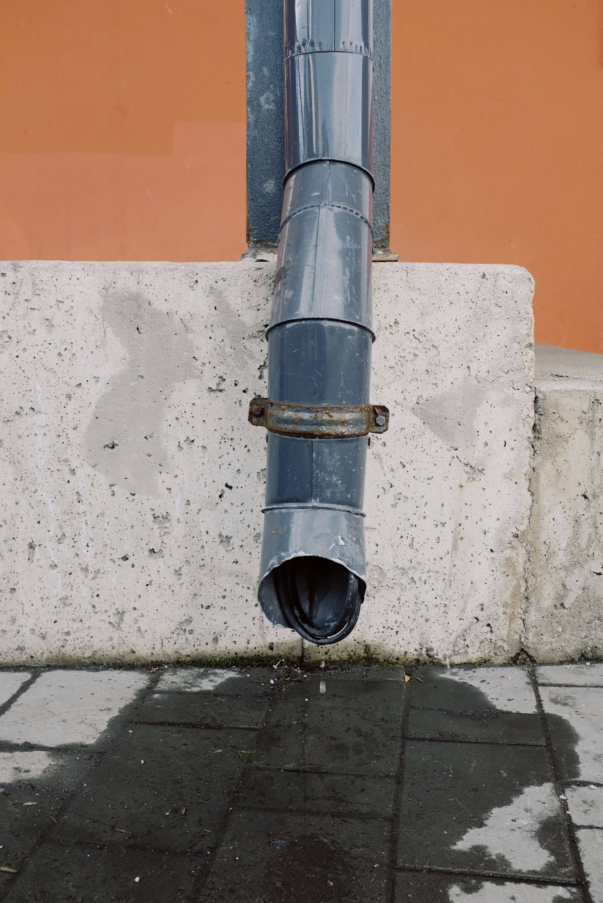 A downpipe discharging water directly onto a sidewalk beside a foundation wall