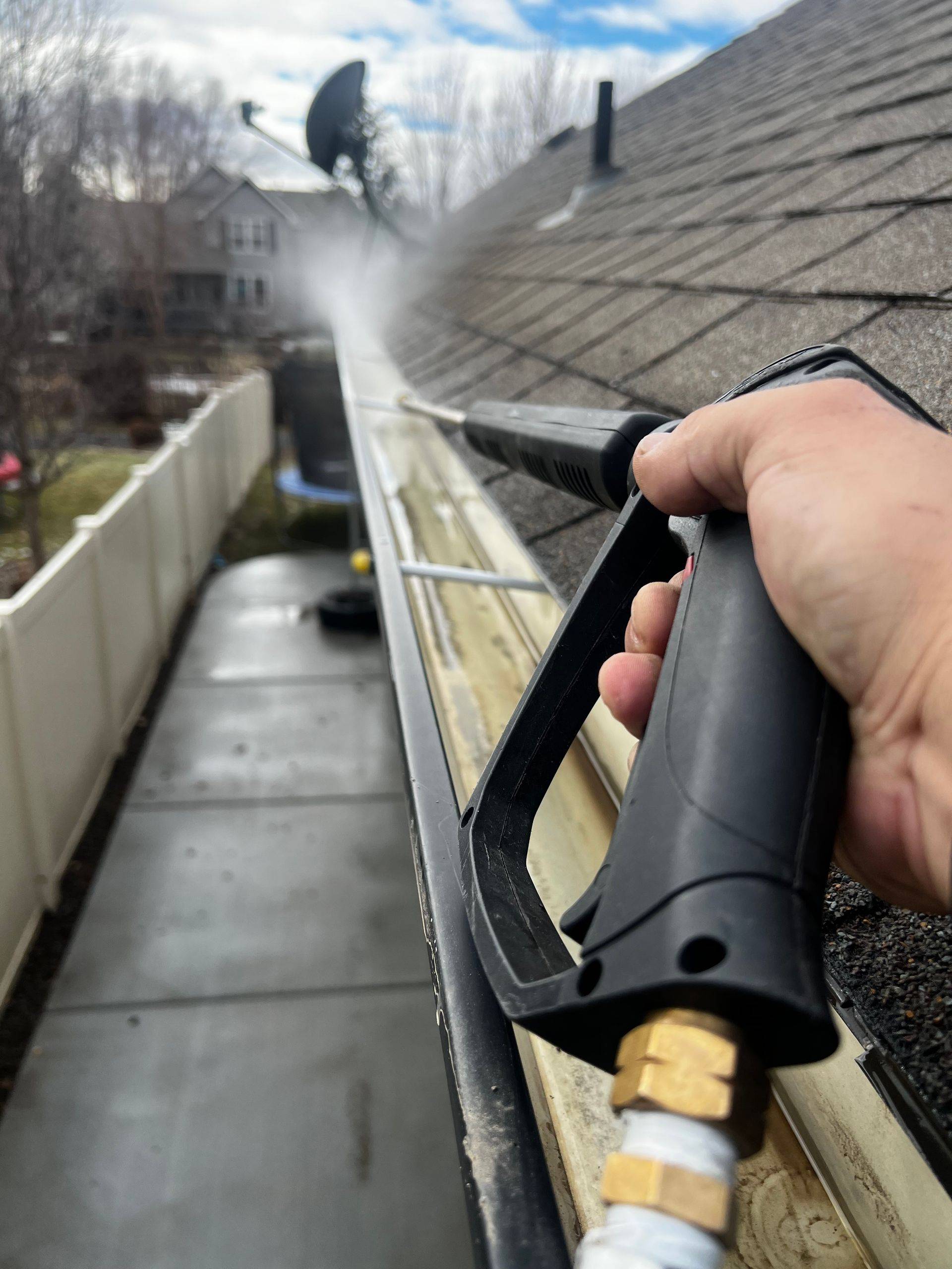 A person clears a gutter at the roofline using a pressure washer wand