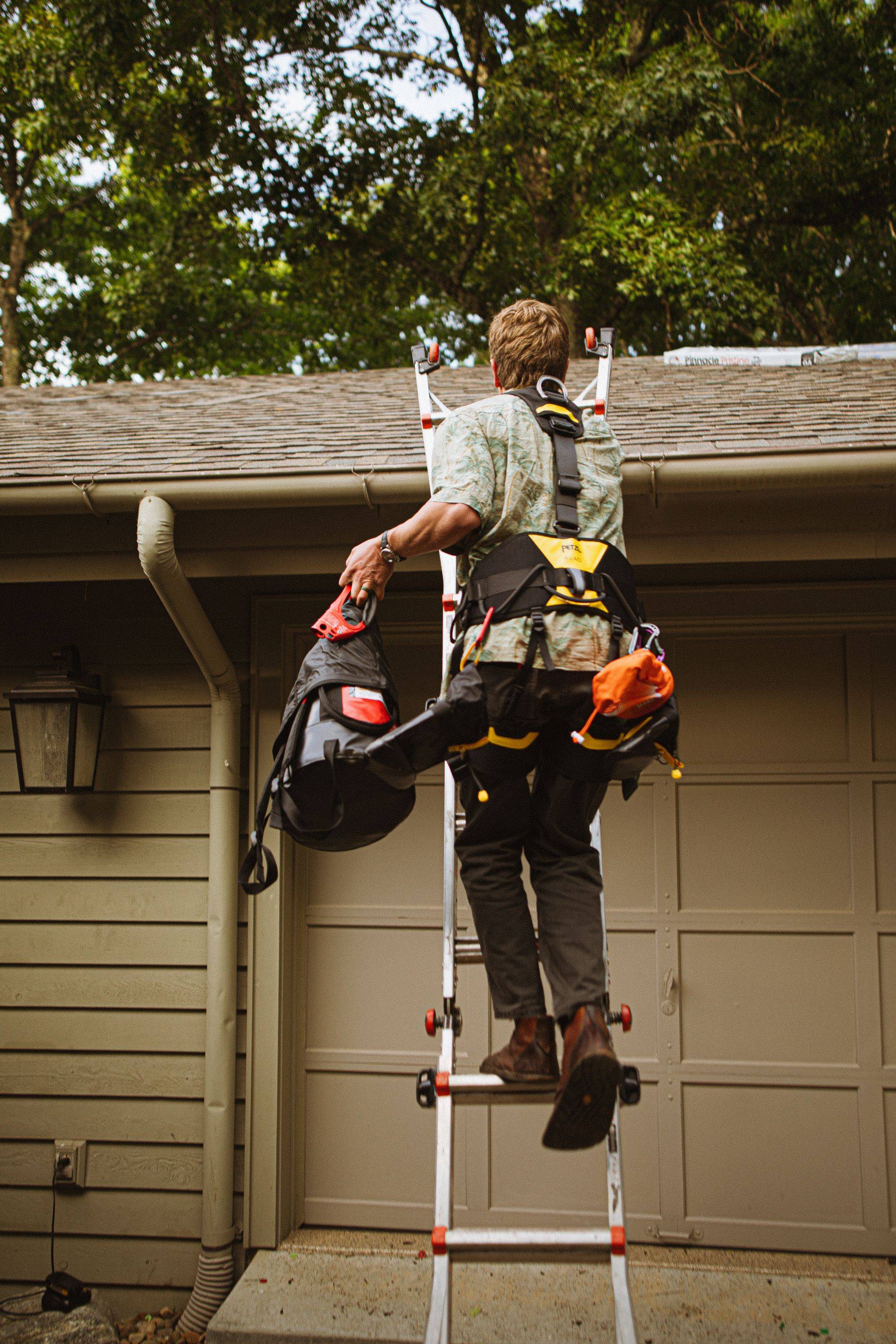 A worker in a full safety harness climbing an extension ladder toward a roof edge